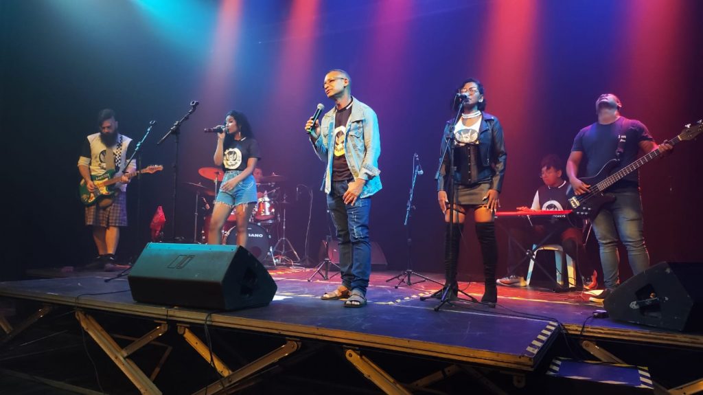 Banda apresenta as canções com o professor John no vocal: alunos e amigos músicos seguram o vocal e o instrumental